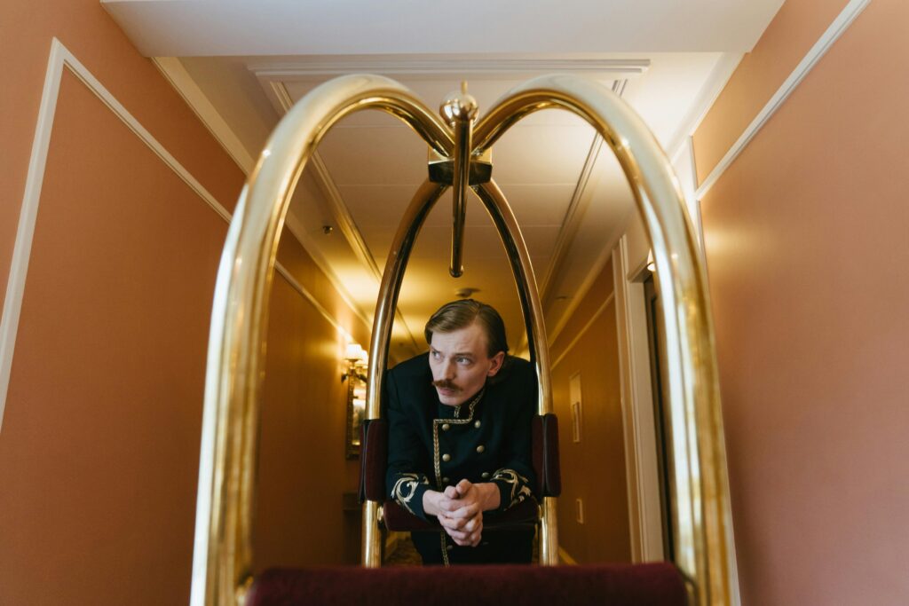 Hotel bellhop dressed in a vintage uniform leaning on a luggage trolley in a brightly lit hallway.